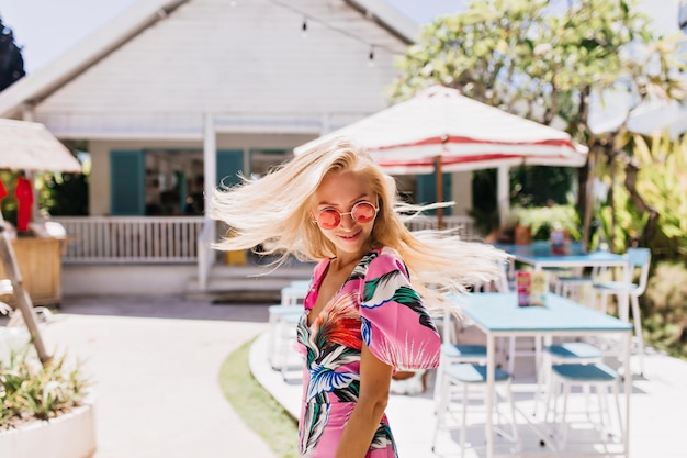 Graceful blonde woman with tanned skin posing near outdoor cafe Portrait of fascinating fairhaired lady in summer dress dancing at resort