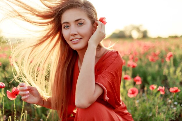 Graceful  blonde woman wraps herself over her shoulder and looking. Windy hairs.  Picturesque poppy field in warm sunset colors.