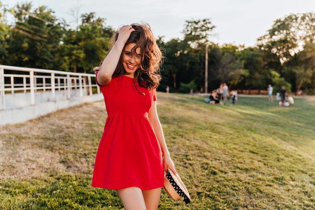 Graceful caucasian lady with brown hair holding straw hat on nature  Gorgeous brunette woman in red dress spending time at field.