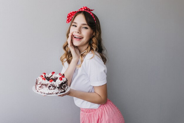 Graceful curly white lady celebrating something with strawberry pie. Portrait of glamorous birthday girl posing with cake.