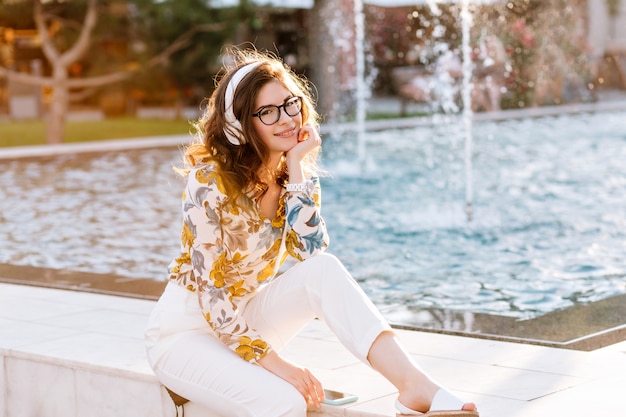 Изящная темноволосая девушка в модных белых штанах отдыхает в парке у красивого фонтана с игривой улыбкой