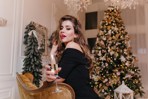 화이트 와인 잔을 들고 장식 된 크리스마스 트리에 대한 포터를 위해 포즈를 취하는 검은 상단의 우아하고 매혹적인 아가씨