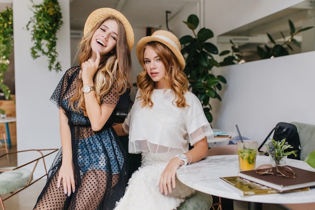 Graceful girl in denim shorts with long shiny hair dancing with smile near friend in elegant white attire