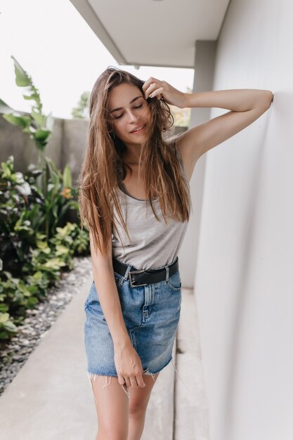 Graceful girl in denim skirt posing with eyes closed near white wall. Outdoor portrait of lovely brunette lady standing near house with bushes