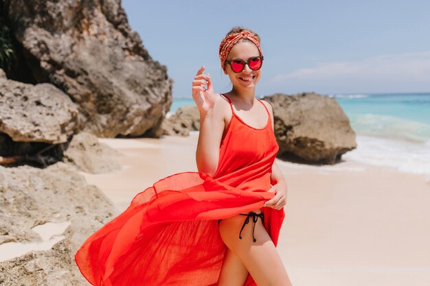 Изящная девушка в красном платье позирует с красивой улыбкой. Фотография стройной загорелой женщины носит ленту, развлекаясь на морском курорте в выходные.