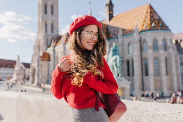 Graceful girl in red outfit looking away with smile, visiting old european castle