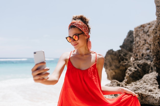 Graceful girl in sparkle sunglasses making selfie in weekend at summer resort. Outdoor shot of blissful tanned lady taking picture of herself while chilling at ocean beach.