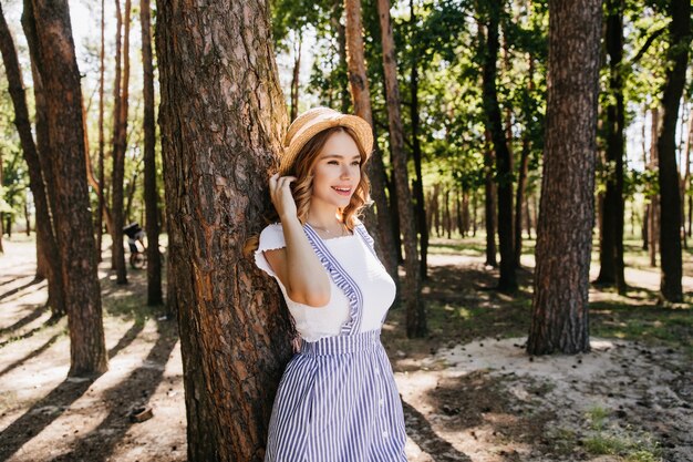 Изящная девушка в соломенной шляпе, наслаждаясь хорошим днем летом. Наружное фото изысканной женской модели, позирующей с улыбкой в лесу.