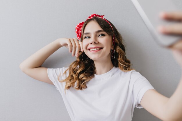 Graceful girl in trendy earrings using smartphone for selfie. Indoor shot of pretty european lady in white attire posing at home.