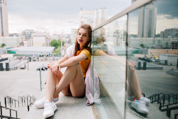 Graceful girl with serious face expression waiting friend on parapet enjoying amazing city view