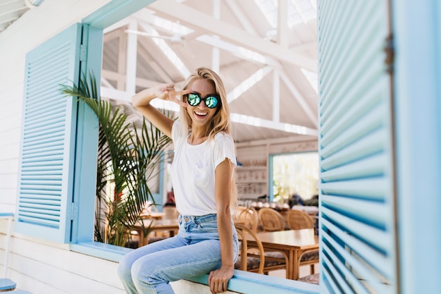 Graceful happy woman sitting on window sill and playing with her blonde hair.