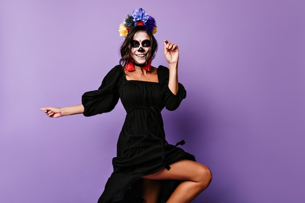 Изящная смеющаяся девушка в черном платье танцует в день мертвых. Радостная латинская дама с макияжем зомби празднует хэллоуин.