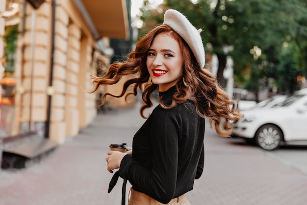 Foto gratuita graziosa ragazza dai capelli lunghi allo zenzero guardando sopra la spalla. donna graziosa di risata in berretto che gode della passeggiata.