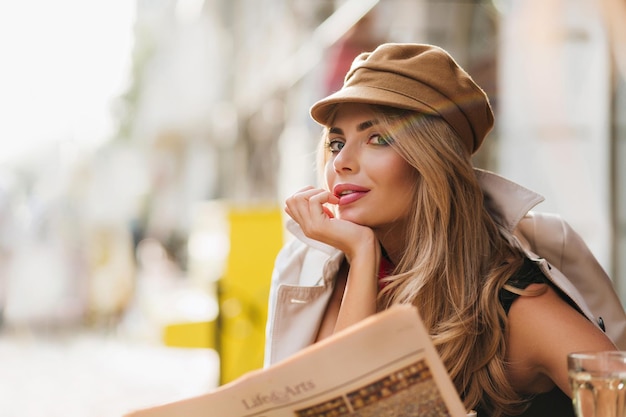 明るいメイクで優雅な長髪の女性は、ぼんやりとした街の背景にふざけてポーズをとって笑っています。ショッピングや新聞を読んだ後に休んでいる茶色の帽子のトレンディな女の子のクローズアップの肖像画。