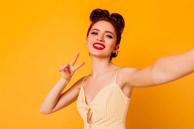 Graceful pinup lady showing peace sign. Studio shot of glad girl with bright makeup taking selfie on yellow space.