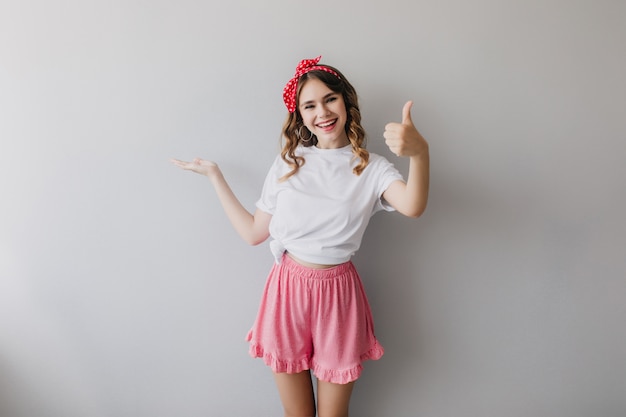 Graceful positive girl in pajamas posing with thumb up. Indoor shot of enthusiastic young lady expressing happiness