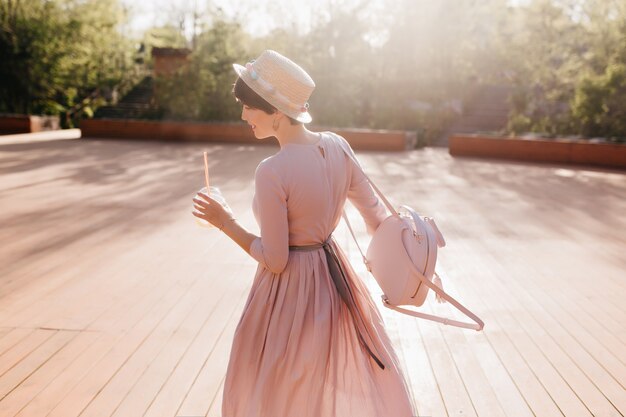 Graceful shapely girl in retro dress dancing outdoor under sunlight, holding trendy backpack