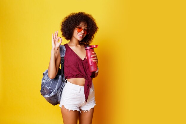 Изящная спортивная черная женщина, стоящая на желтом и держащая розовую бутылку воды. В стильной летней одежде и рюкзаке.