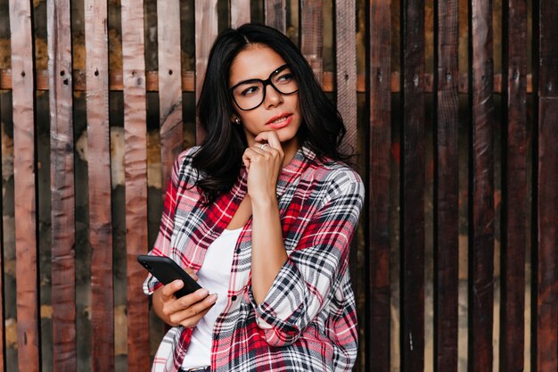 Graceful tanned woman in glasses standing on wooden wall. Outdoor shot of relaxed brunette girl dreamy posing with phone.