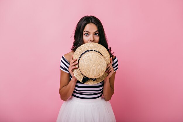 Graceful tanned woman hiding face beside straw hat. Indoor portrait of adorable latin lady playfully posing.