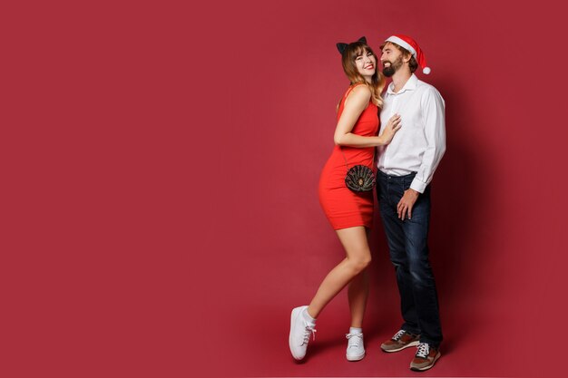 Graceful woman in cat ears masquerade hat and red short dress with her boyfriend posing on red. New year party. Full length .