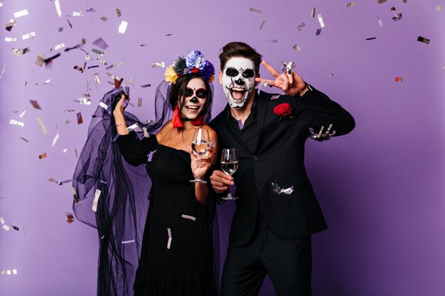 Graceful woman in colorful wreath celebrating halloween with boyfriend. Couple of vampires drinking champagne.