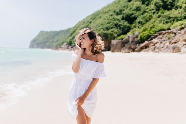 밝은 미소로 바다를 바라 보는 드레스에 우아한 여자. 열대 섬에서 시간을 보내는 화려한 금발 아가씨의 야외 사진.