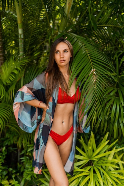 Graceful Woman model in red swim wear with long straight hair posing in tropical nature Perfect tan body
