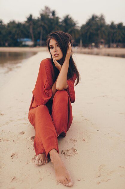 Graceful woman posing on the beach, sitting on sand in red dress