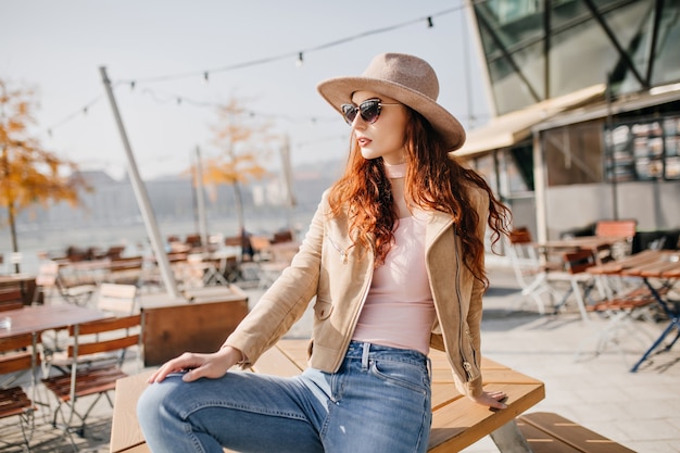 Graceful woman with long ginger hair looking around in outdoor cafe in autumn weekend