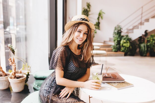 Graceful young lady resting at the table with glass of cold tea and magazines