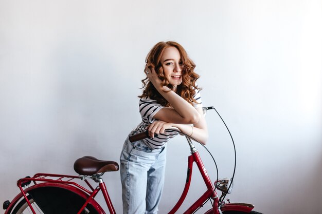 Graceful young lady with red hair standing near her bicycle during photoshoot. Indoor portrait of curly girl in blue jeans isolated.