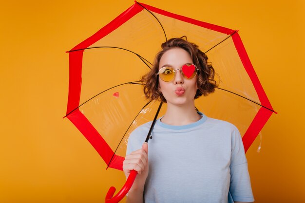 Graceful young woman in blue shirt posing with kissing face expression. Studio shot of pretty female model with curly hair fooling around during photoshoot with umbrella.