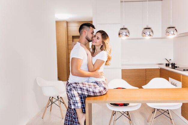 Graceful young woman in pajama kissing husband in cozy kitchen. Winsome girl sitting on table and looking at boyfriend with smile.