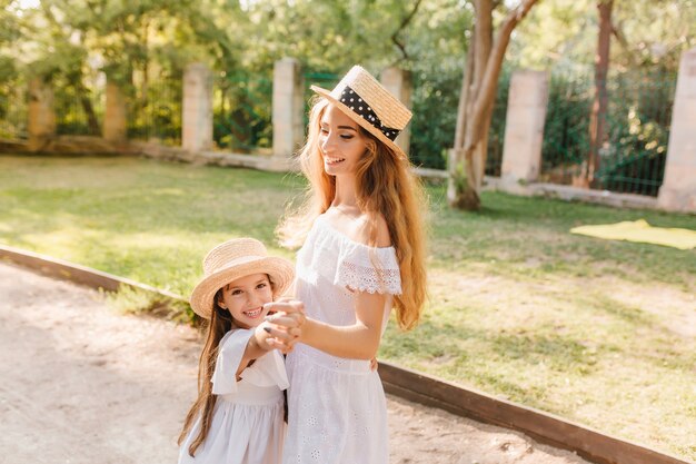 Graceful young woman in white dress dancing with daughter on the alley and smiling. Outdoor portrait of charming mom in straw boater holding hands with joyful child wanted to play.