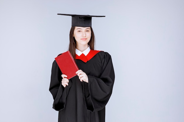 Studente laureato che indossa mortarboard che tiene libro rosso e posa. foto di alta qualità