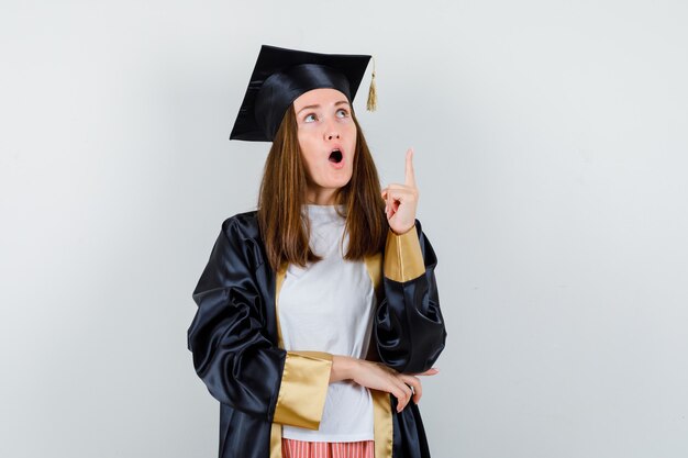 Graduate woman pointing up in casual clothes, uniform and looking amazed. front view.