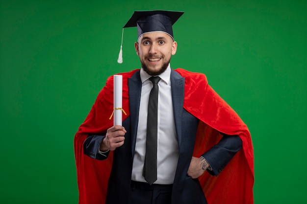 緑の壁の上に立って幸せで陽気な笑顔の卒業証書を保持している赤いマントの卒業男