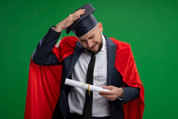 Graduated man in red cape holding diploma looking confused with hand on his head for mistake standing over green wall