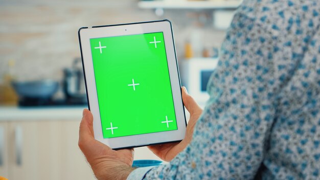 Grandgather using tablet pc with green screen in kitchen while enjoying his breakfast. Elderly person with chroma key isolated mock-up mockup for easy replacement