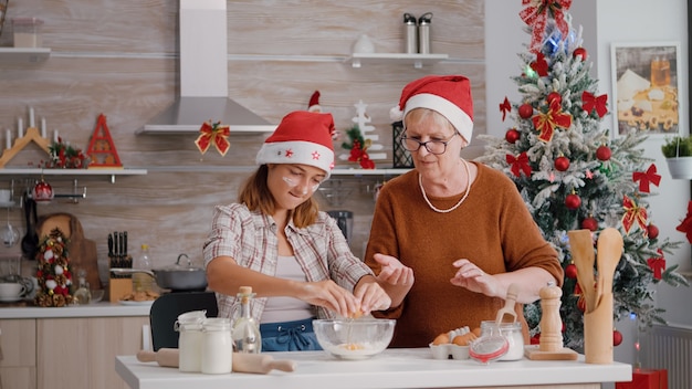 Grandma breaking egg helping grandchild preparing festive cookie dough in culinary kitchen