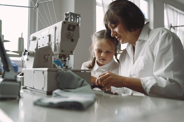 Grandma with little granddaughter sew clothes in the factory