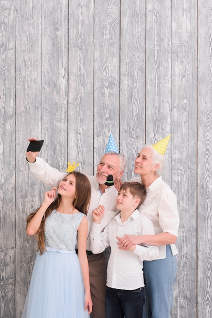 Grandparent wearing party hat taking selfie on mobile phone with their grandchildren holding paper props