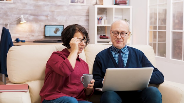 ノート​パソコン​で​の​ビデオ​通話中​に​家族​と​話している​ソファ​に​座っている​眼鏡​を​かけた​祖父母​。