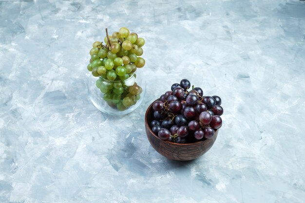 Грозди винограда в глиняной миске и стеклянном горшке под высоким углом на шероховатом сером фоне