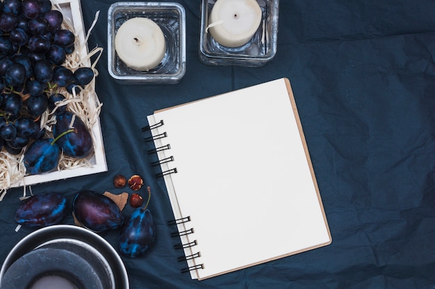 Free photo grapes in crate with plums; candles and blank spiral notepad on tablecloth