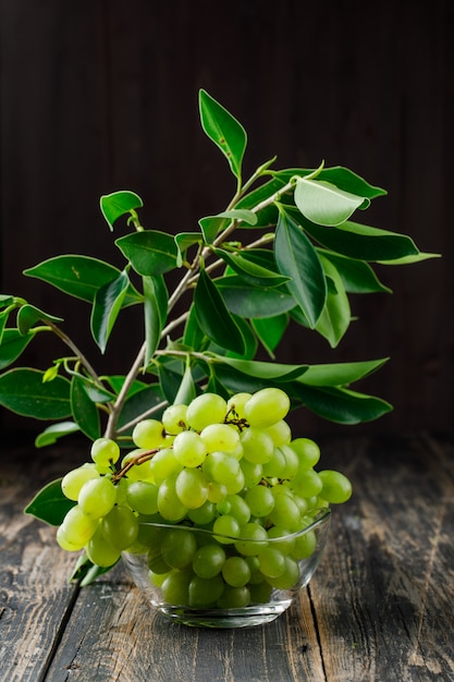 Grapes with leaves on branch in a glass bowl on wooden surface, side view.
