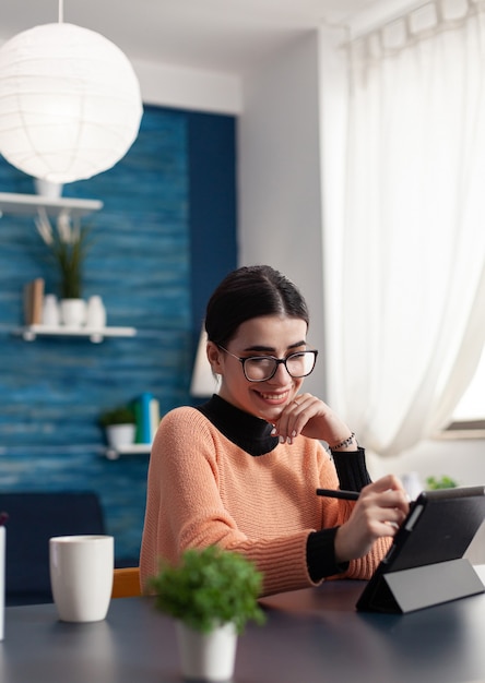 Graphic designer working on creative illustration using graphic digital tablet holding drawer pen in hand retouching draw. Focused student sitting at desk table in living room