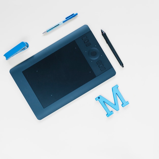 Graphic digital tablet; pen; stapler and m alphabet on white surface