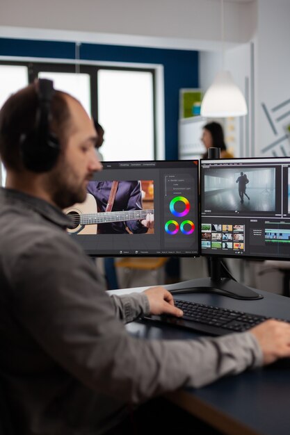 創造的な職場に座ってビデオとオーディオの映像を編集する2つのディスプレイを備えたPCで動作するグラフィックビデオ制作